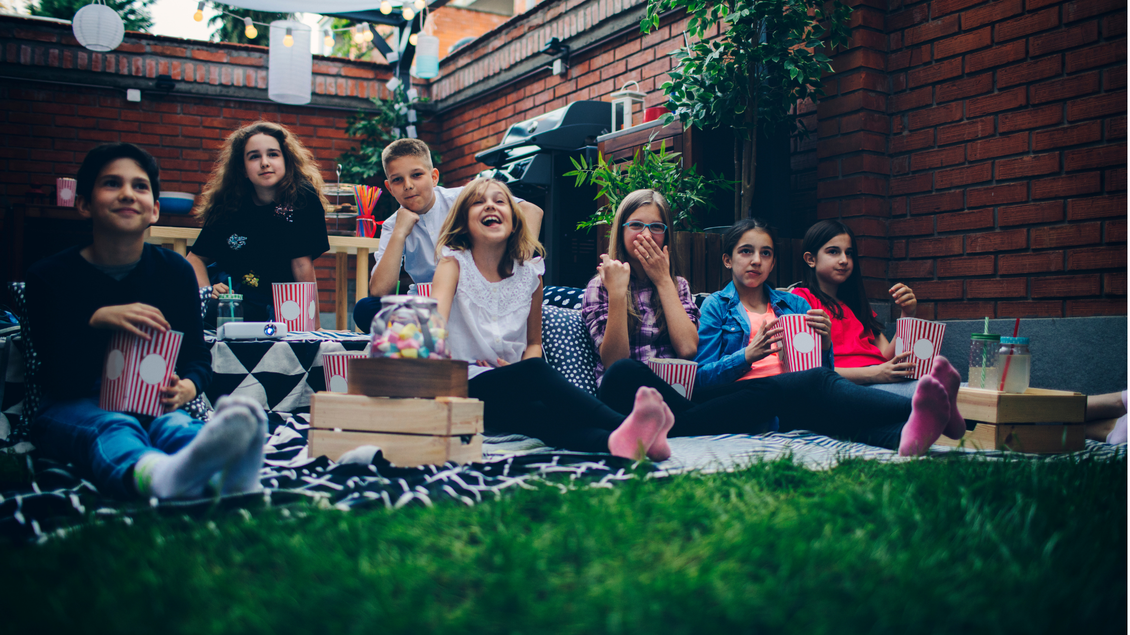 Kids sitting on a blanket on the lawn enjoying popcorn and soda while watching a movie