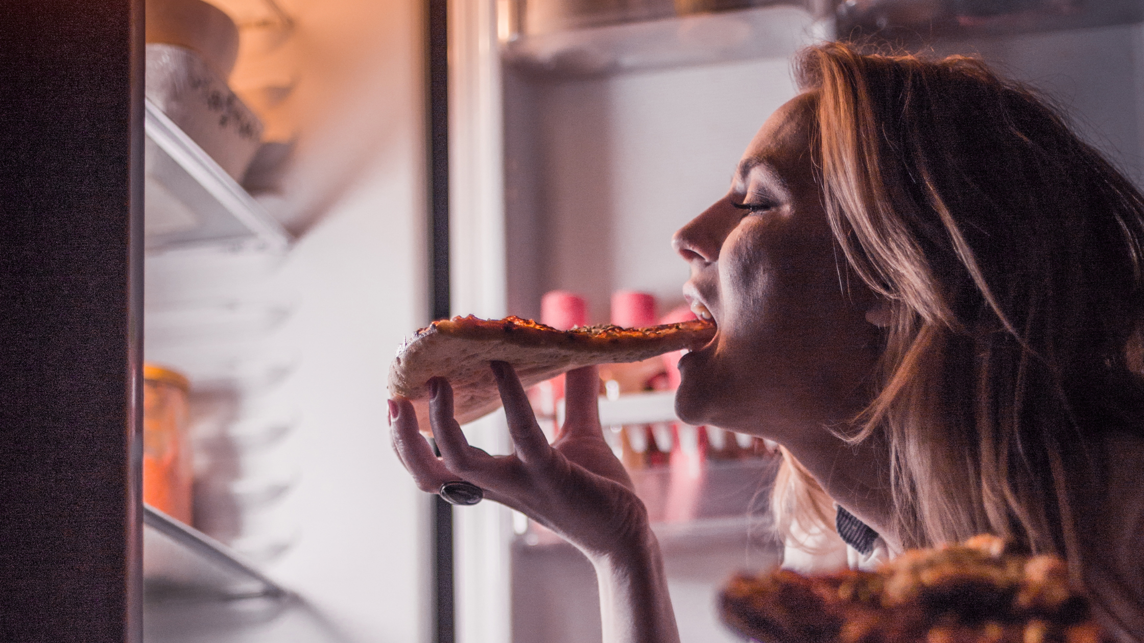 woman eating a slice of pizza out of the fridge in the dark