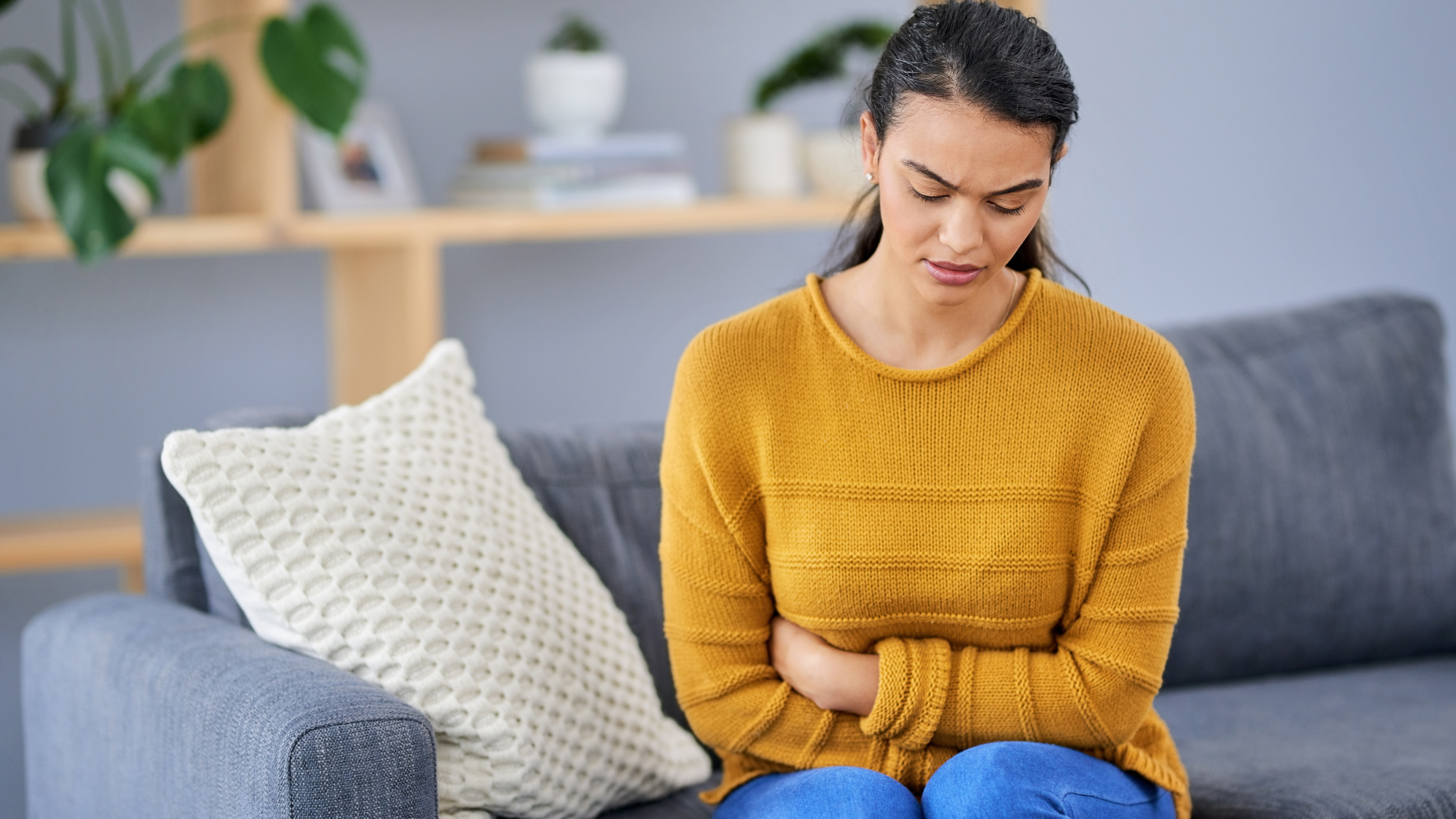 young woman sits on a couch, clutching her abdomen