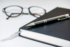 A pen resting on a journal, with a pair of glasses in the background
