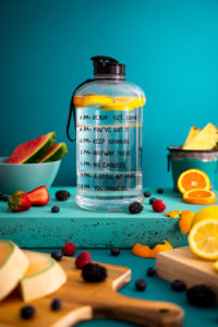 Various citrus and berries in front of a bottle of water with lemon slices floating in it