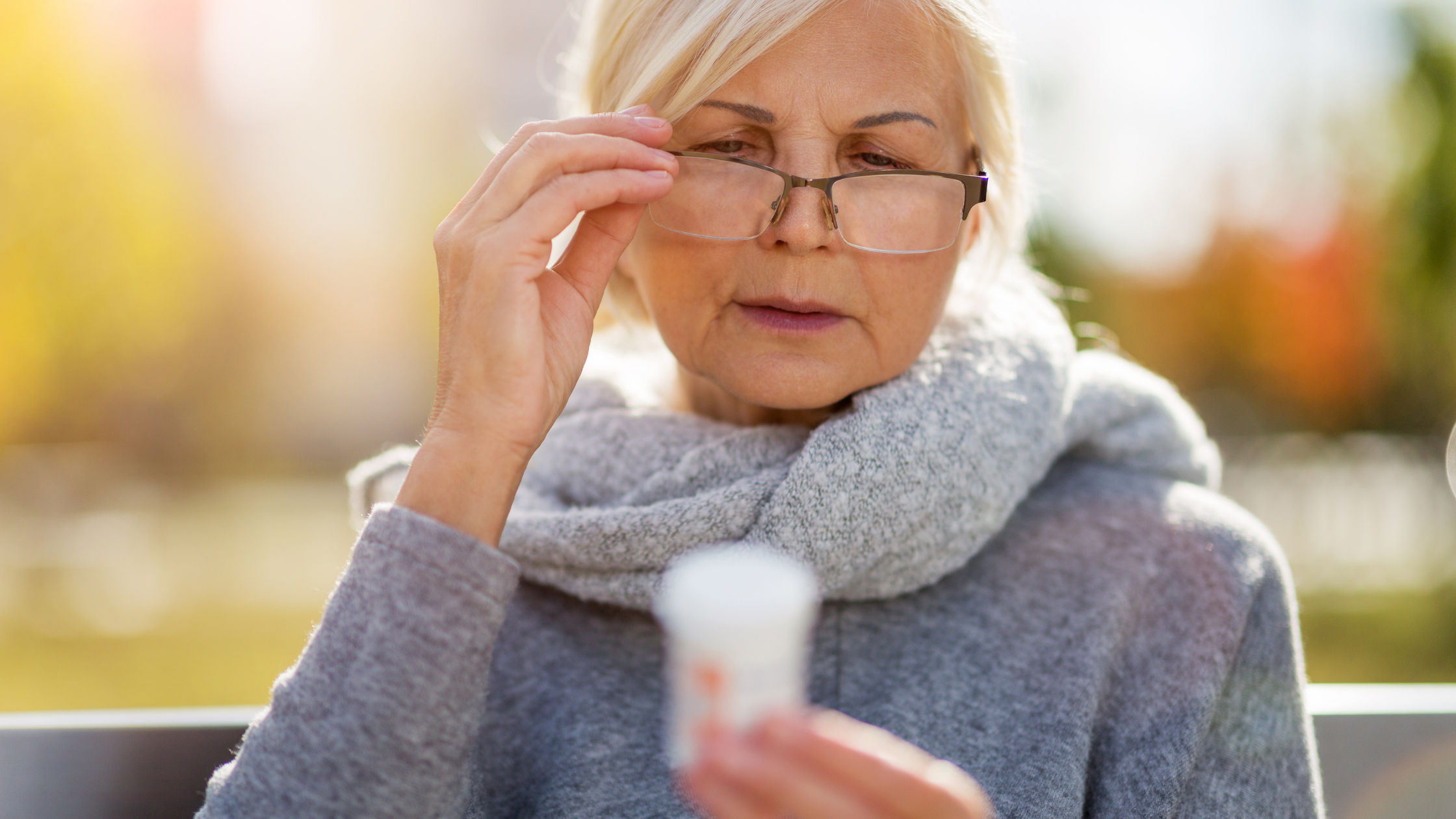 Older woman in the park adjusts her glasses to read held medication bottle