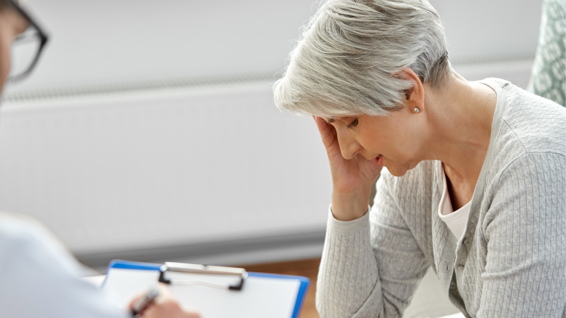 senior woman holds her head in her hand while a medical professional listens with a clipboard