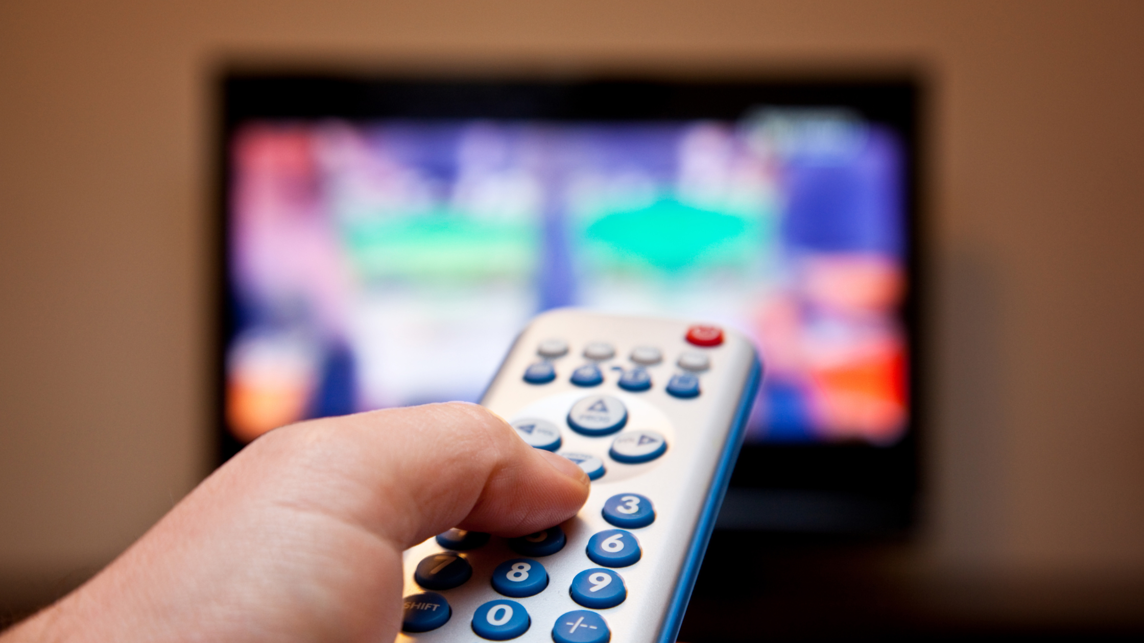 adult hand holds a TV remote in front of a blurry screen