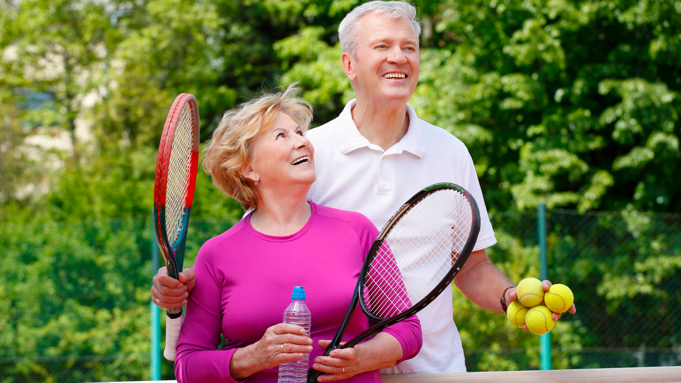 Smiling older couple on a tennis court holding rackets and balls