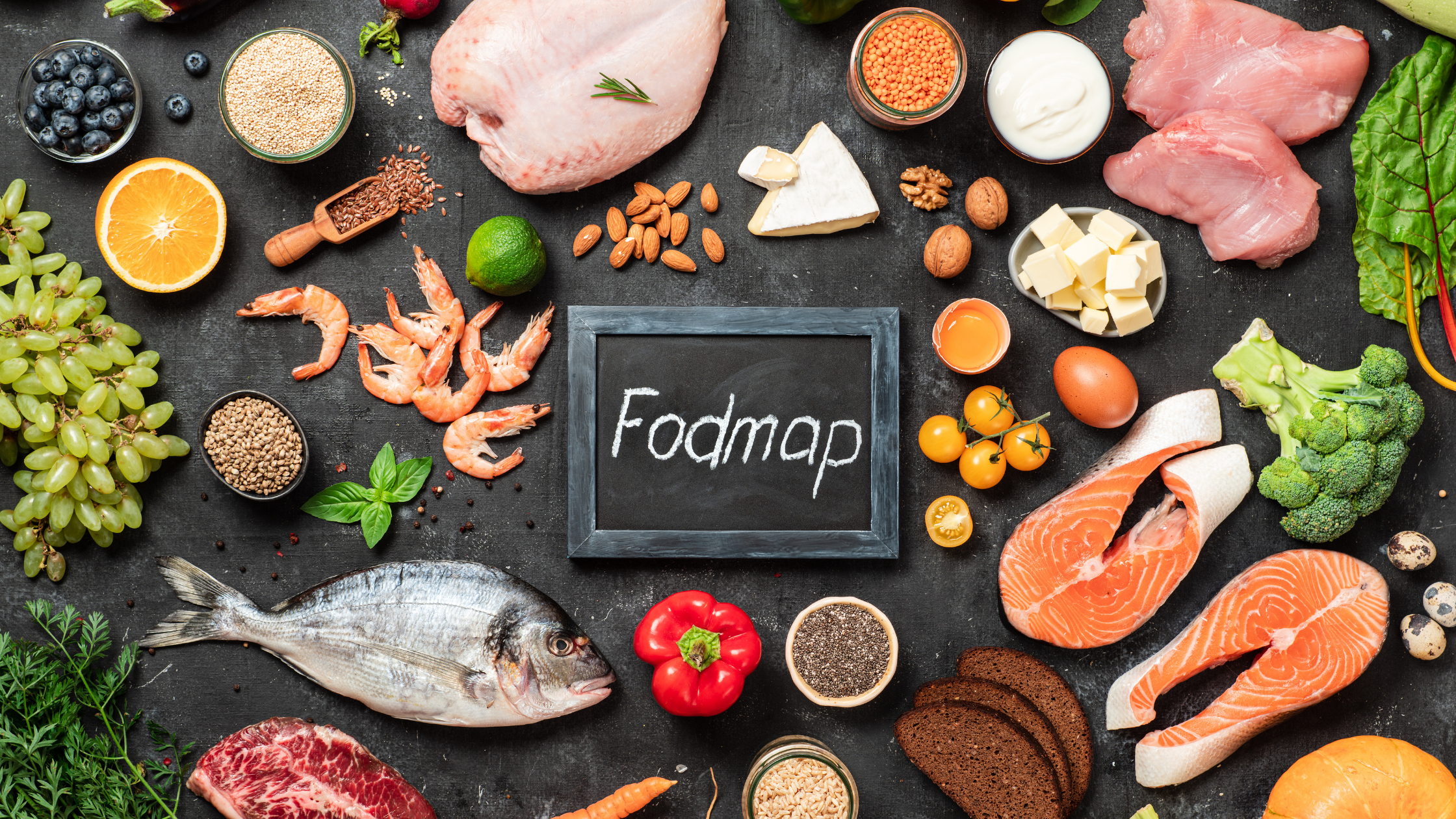 The word "Fodmap" written in chalk on a slate board surrounded by various healthy-looking foods, sliced oranges, cherry tomatoes, shrimp, salmon, broccoli, cheese, nuts, herbs