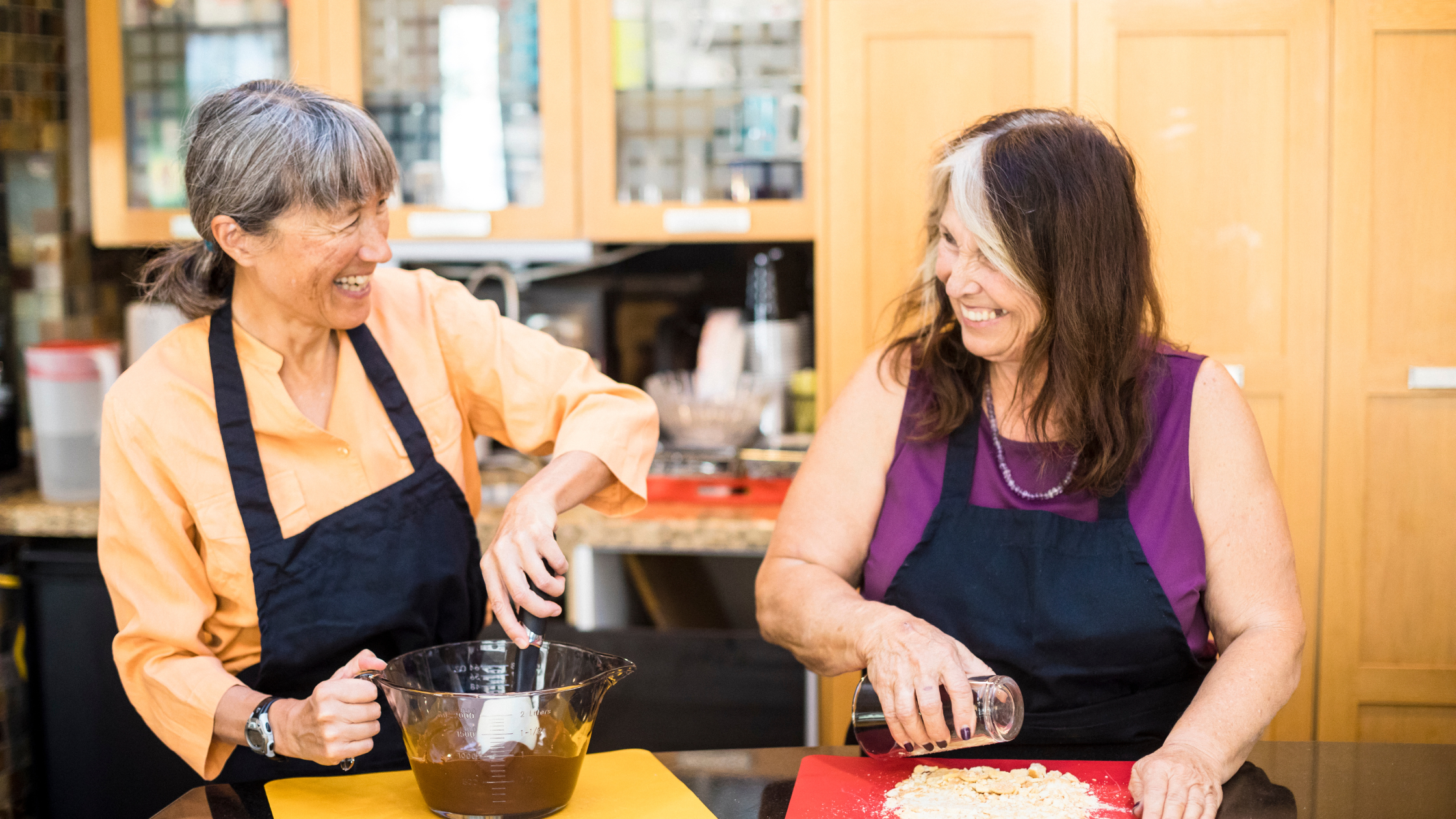 two senior women smile at each other while they cook separately 