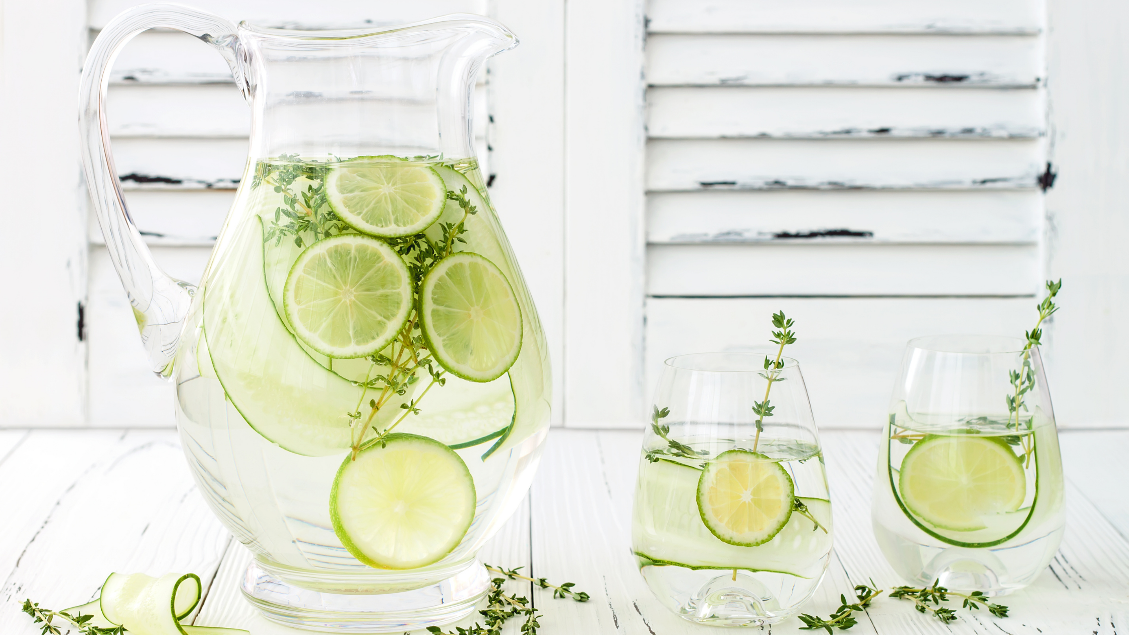 Glass pitcher of water with lime slices, sprig of thyme on a white wooden table