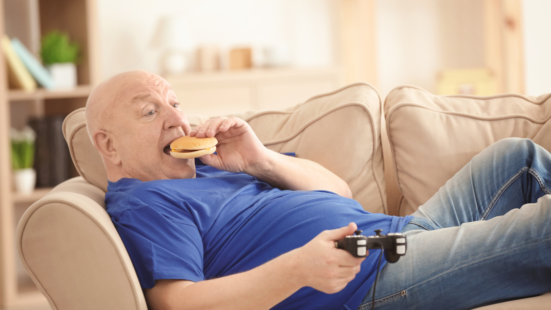 senior man reclining on a couch stuffs a burger in his mouth 