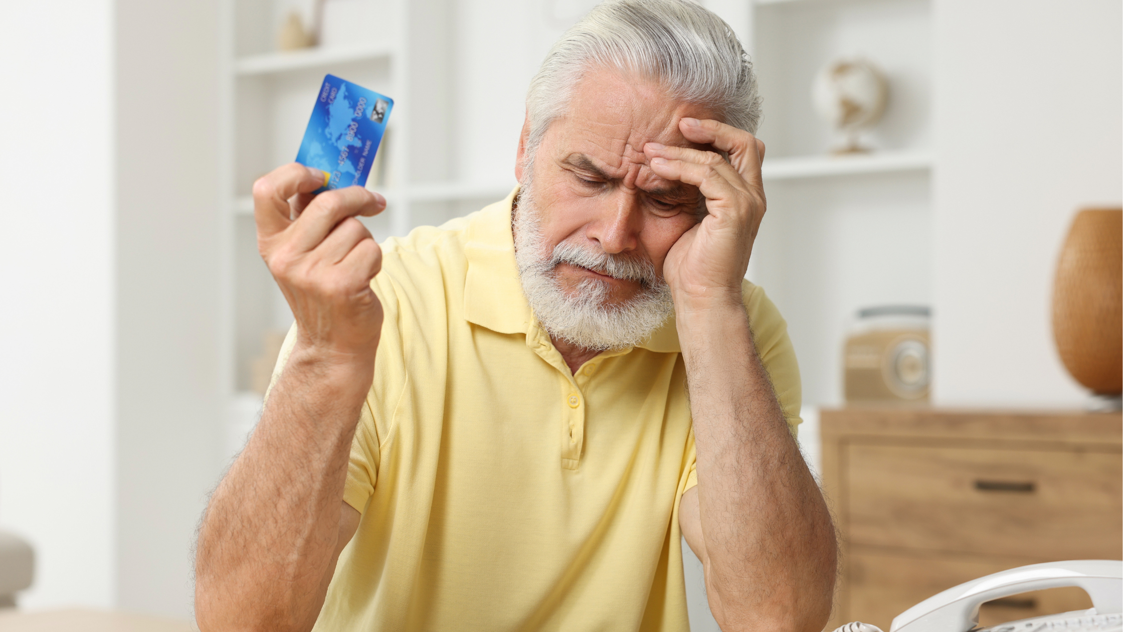 concerned-looking senior man holds head in one hand, a credit card in the other