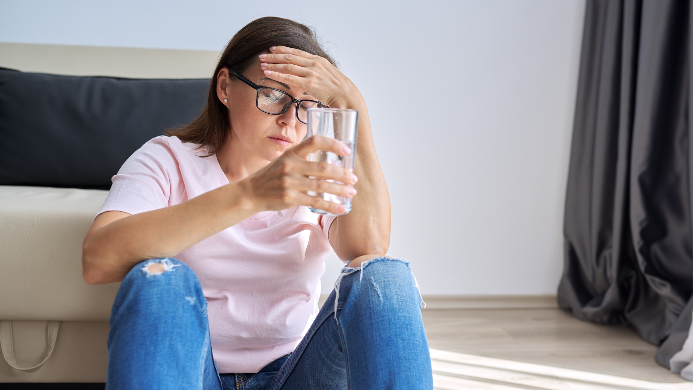 woman sits on the floor with a glass of water and her hand on her forehead