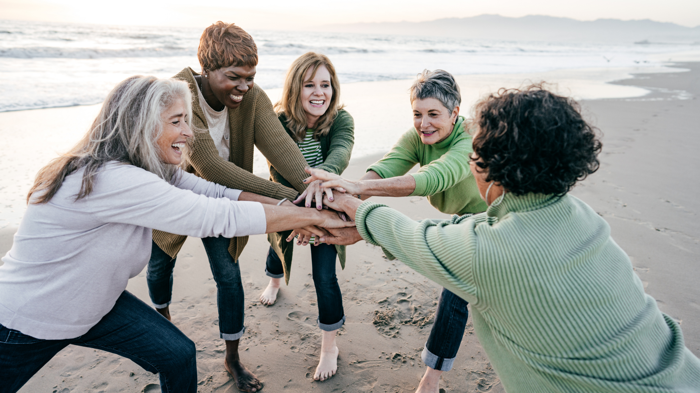 A happy group of women on the beach in a circle with their hands placed one over the other in the middle about to do a group cheer