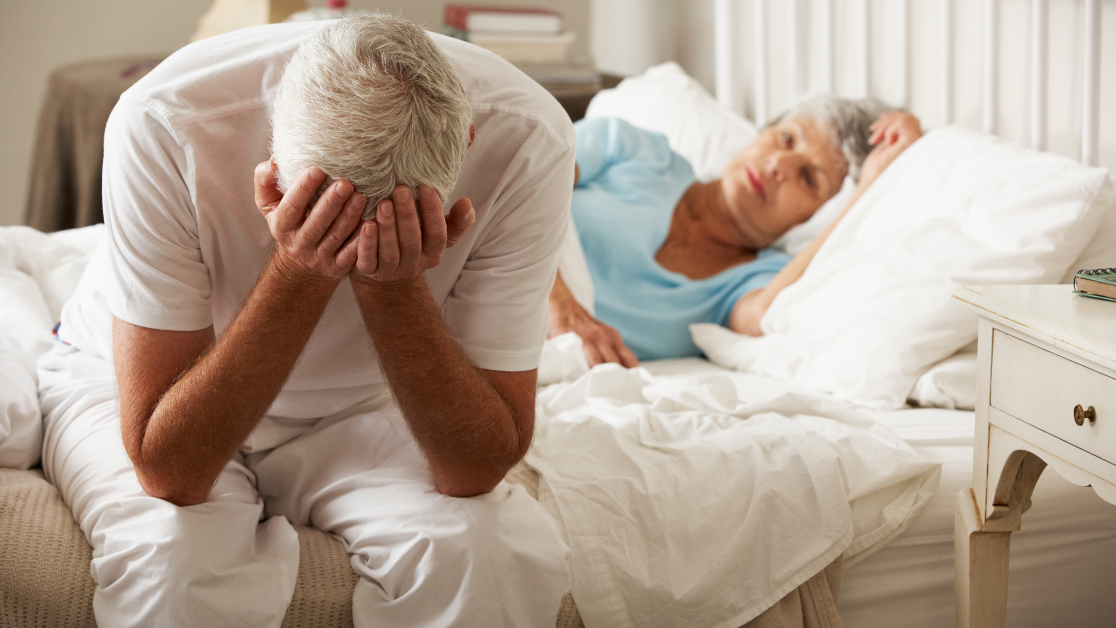 man sits on the side of a ben with his head in his hands, with a sad woman laying behind him