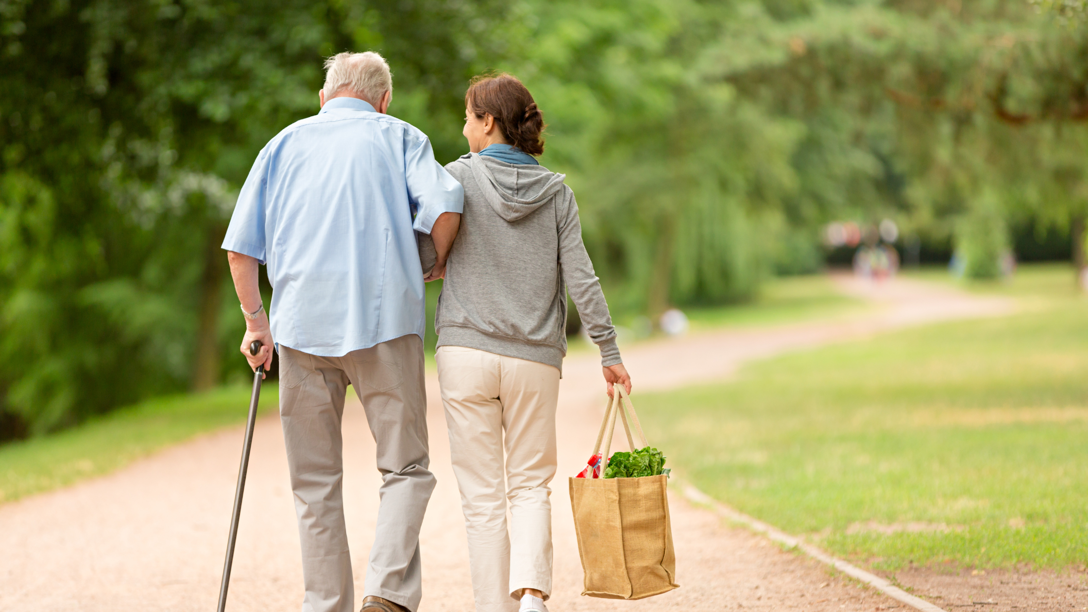 senior man walks beside a smiling younger woman carrying his groceries