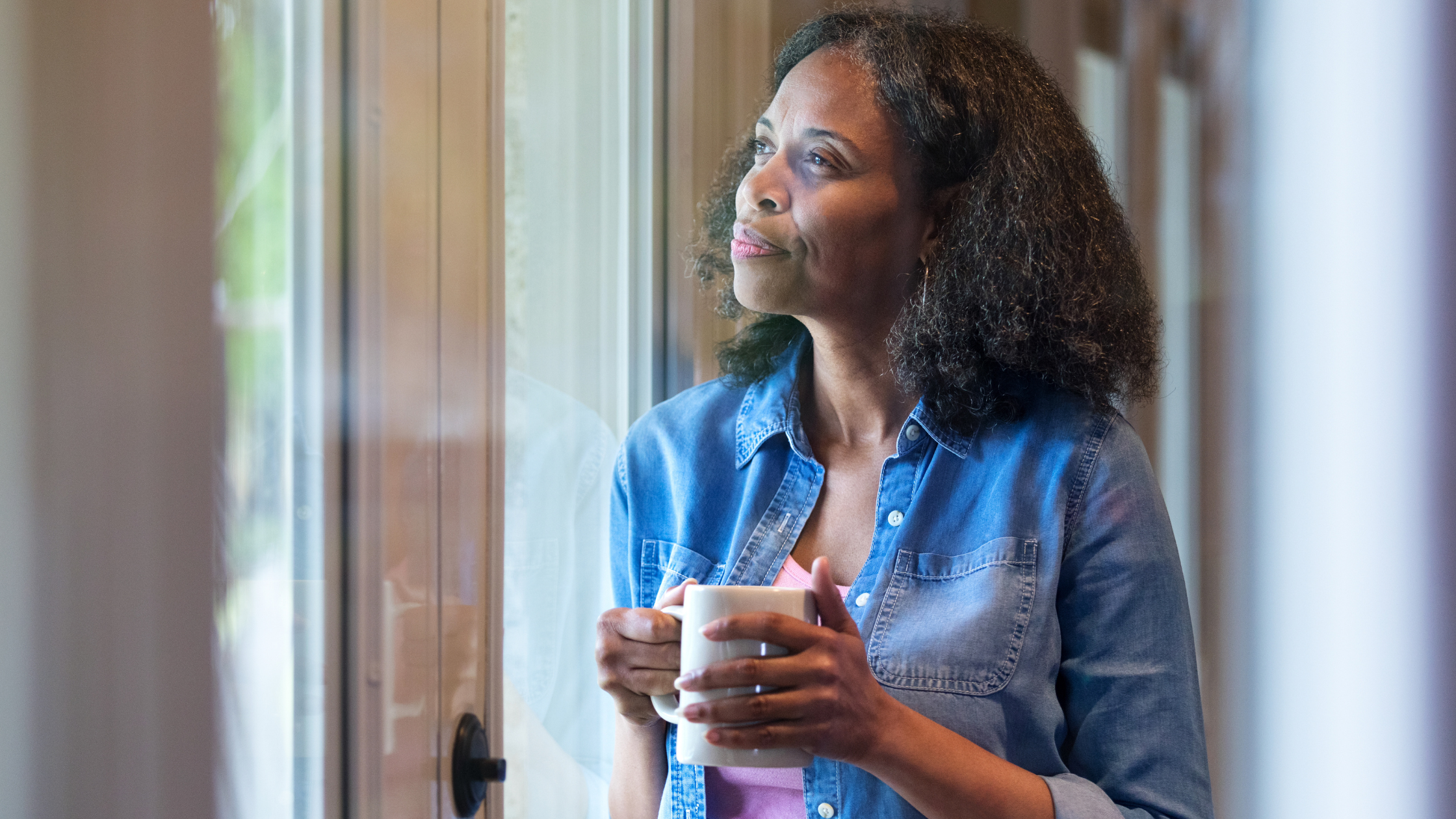 woman holding a warm beverage in a mug stares hopefully out a window