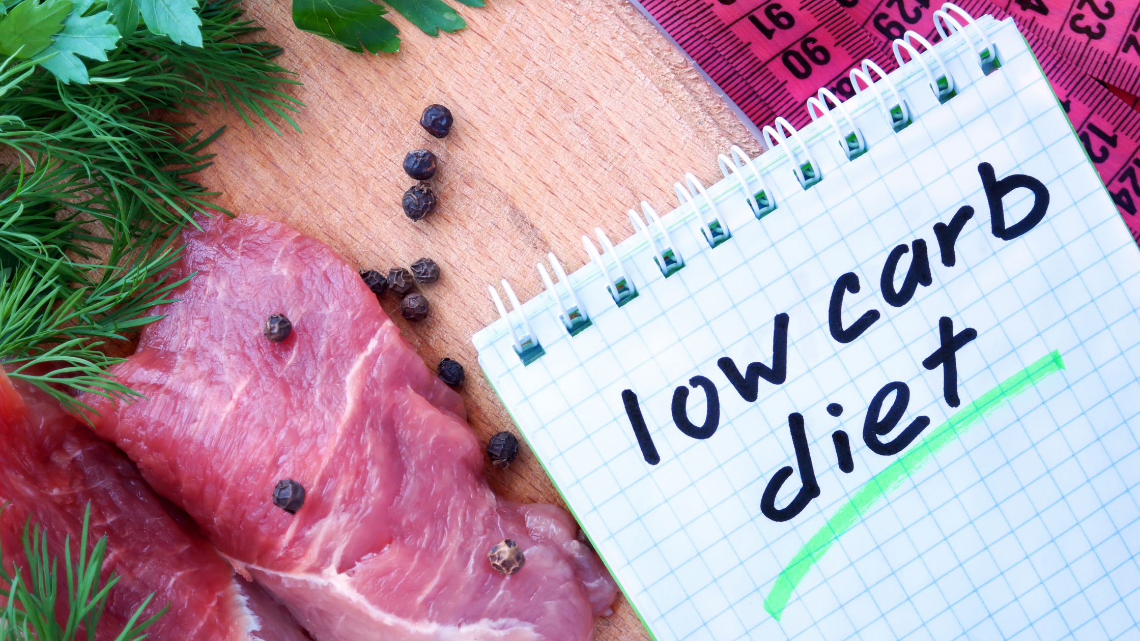 A notepad with the words "Low Carb Diet" written on it lying on a wooden table next to sliced meat, berries, and herbs