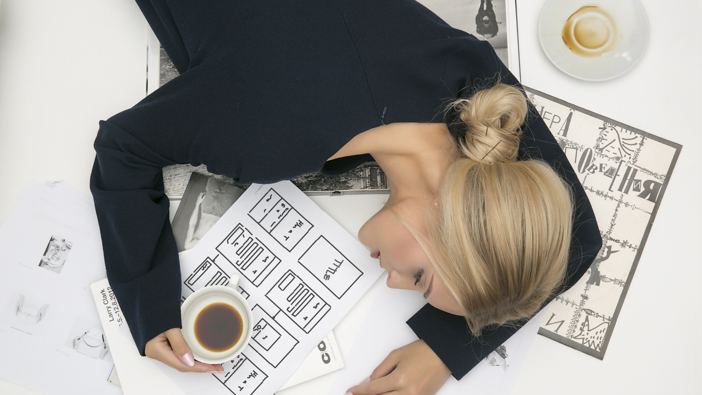 Woman asleep on a table full of graphs, with a cup of coffee in her hand