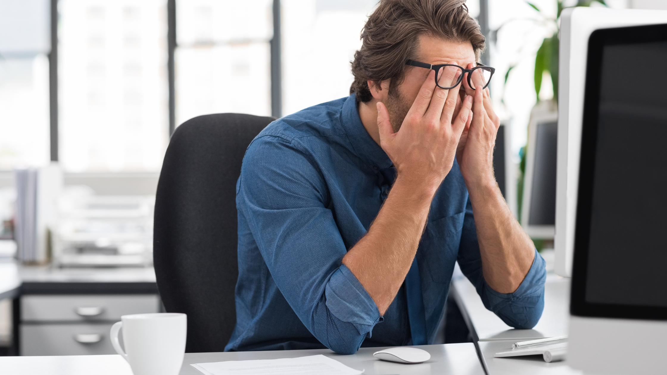 man sits in front of a desktop computer with his face in his hands