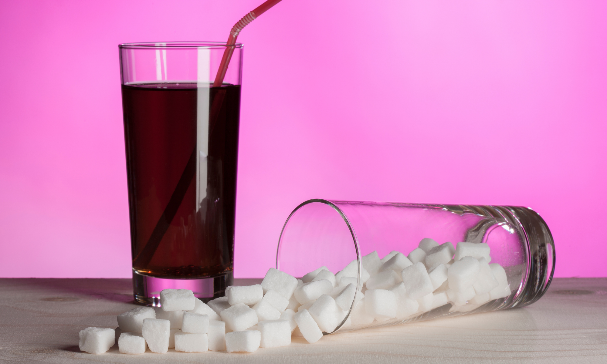 A glass of soda sitting next to a tipped over glass with sugar cubes spilling out of it