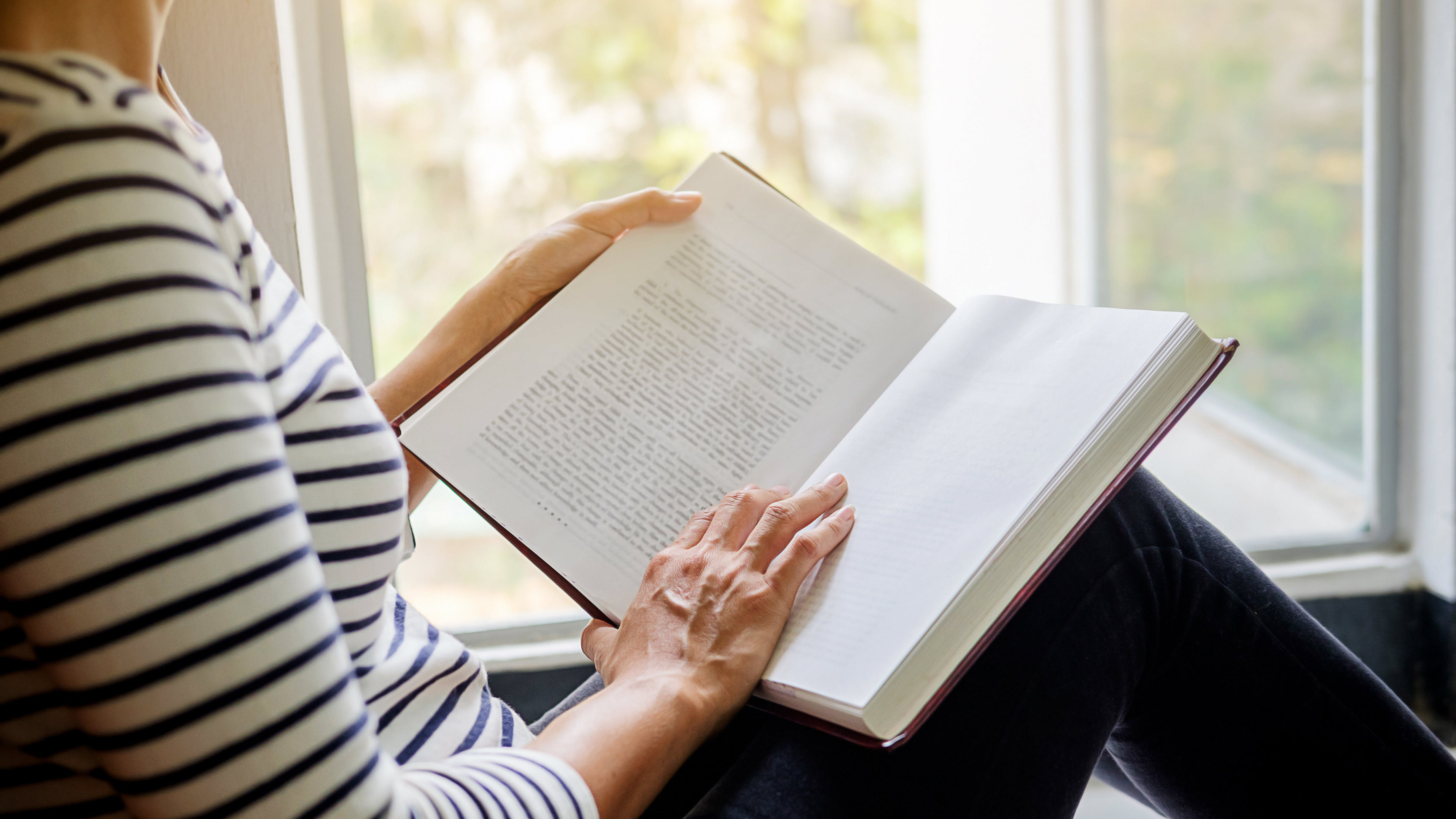 someone sits in a window seat with an open book in their lap