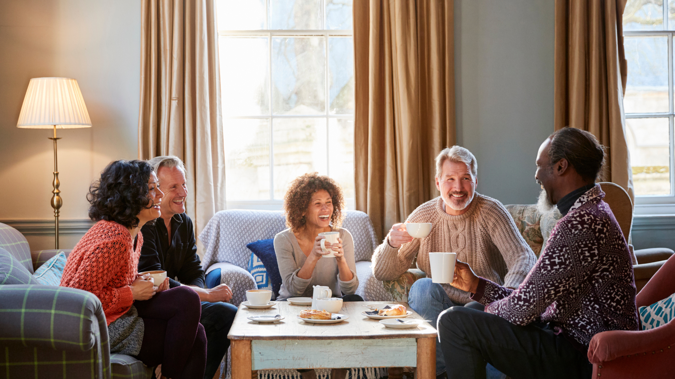 group of middle aged and senior adults sit in a living room drinking from mugs